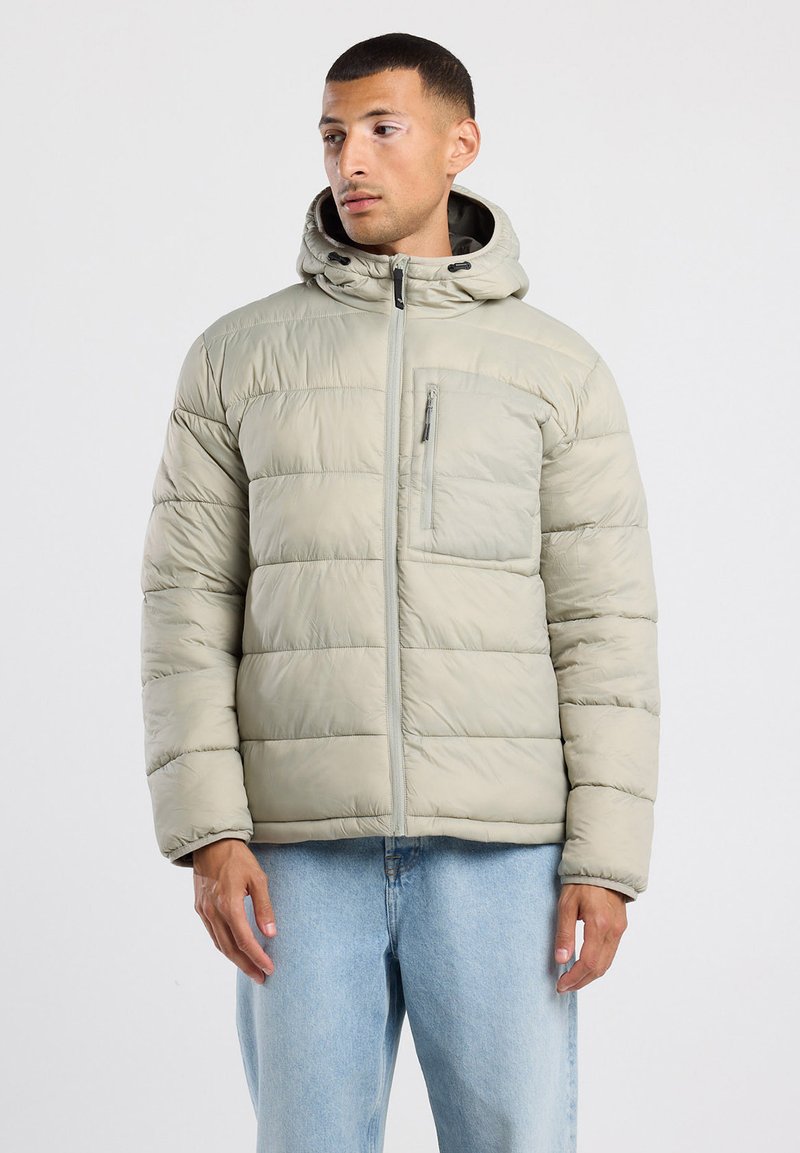 Chaqueta de plumón gris claro con capucha, costuras verticales, bolsillo en el pecho con cremallera y puños elásticos. Lleva puesta sobre unos jeans azules.