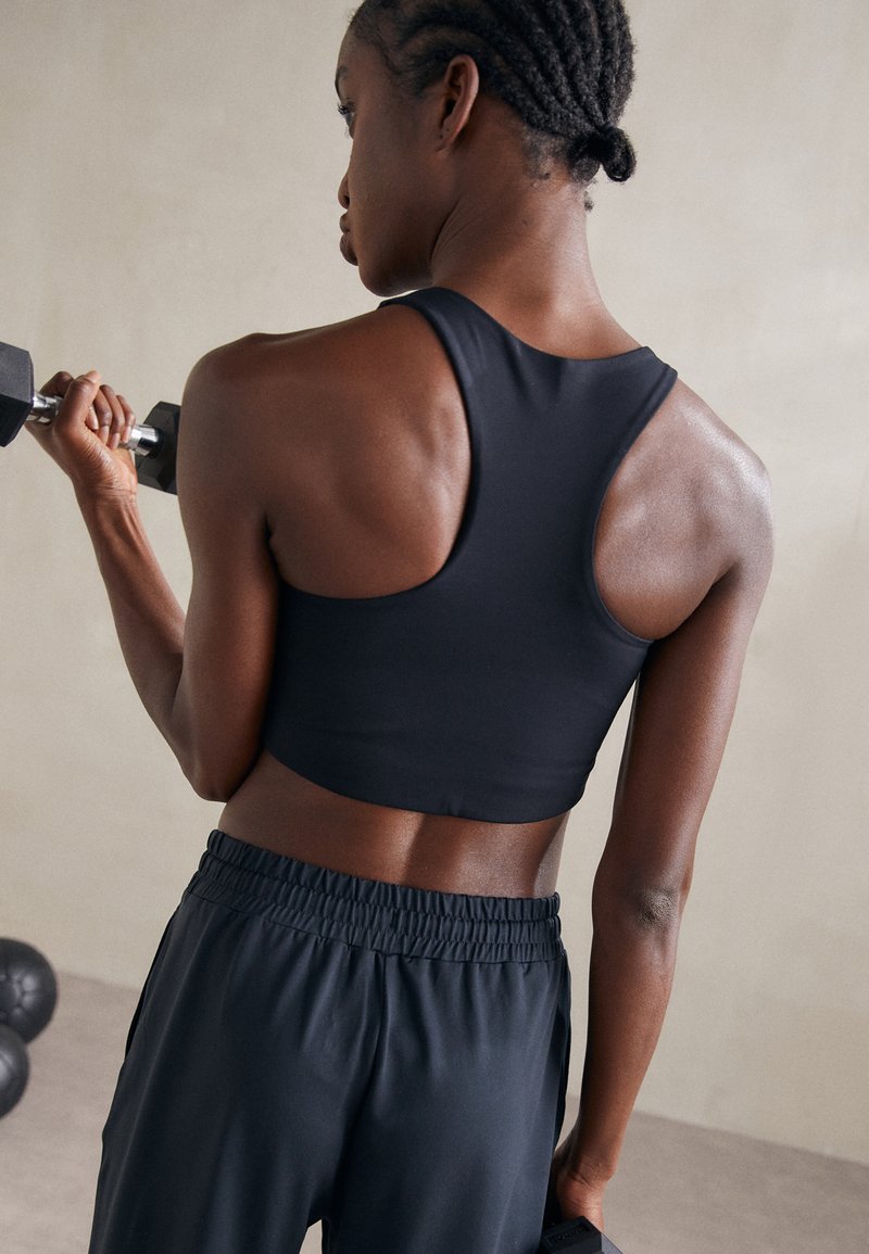 Svart, ermeløs cropped treningsgenser med racerback-design, kombinert med løse svarte treningbukser. Lys glans på den glatte stoffet.