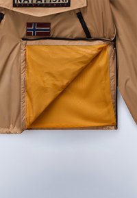 Beige jakke indvendig med en lynlåset mesh-lomme. Inkluderer et lille mærke med et flagdesign i rød, hvid og blå.