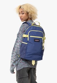 Zaino blu navy con accenti gialli, realizzato in tessuto resistente. Caratterizzato da una tasca anteriore con zip e una tasca laterale in rete, che mostra un design moderno.