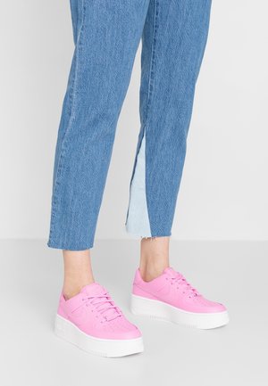 Personne portant un jean court bleu avec des fentes latérales et des baskets à plateforme roses, debout sur un sol gris.