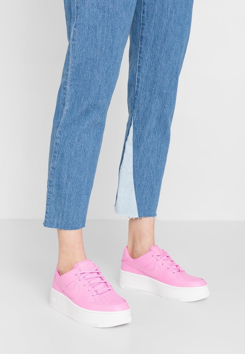 Personne portant un jean court bleu avec des fentes latérales et des baskets à plateforme roses, debout sur un sol gris.