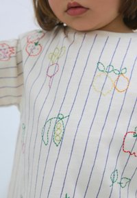 Hvid stof t-shirt med lodrette blå striber, der har farverige broderede frugt- og grøntsagsmotiver i rød, grøn, gul og pink.