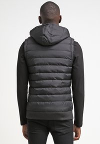 Jachetă neagră, pufosă, fără mâneci, cu glugă și tiv ribbed. Are un exterior neted și cusături verticale. Pur tată peste un tricou negru cu mânecă lungă.