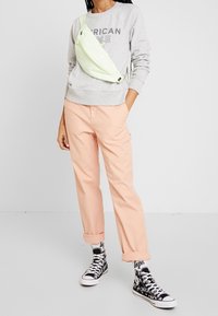 Jasnoszara bluza z nadrukiem, pastelowe brzoskwiniowe spodnie, neonowozielona torba na ramię oraz czarne sneakersy za kostkę z graficznymi skarpetkami.