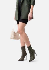 Stivali alla caviglia verdi con punta affilata, tacco a stiletto alto e design morbido; abbinati a una borsa beige testurizzata e pantaloni corti neri.