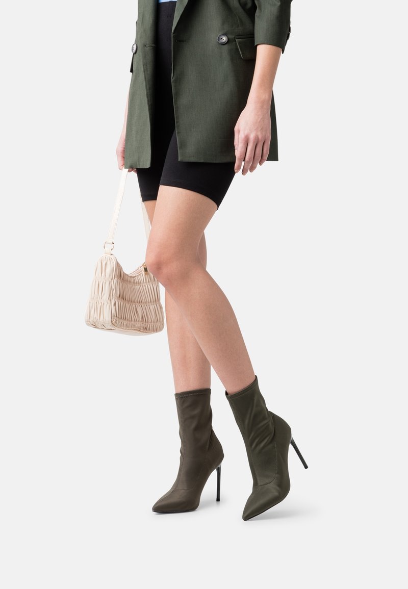 Bottines vertes à orteils pointus, talons aiguilles hauts et design décontracté ; associées à un sac à main beige texturé et un short noir.
