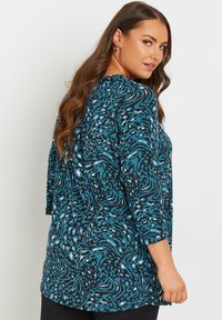 Tunic a maniche lunghe in un motivo astratto blu petrolio e nero. Tessuto morbido e stretchy con una vestibilità ampia e orlo curvo. La vista posteriore mostra maniche a 3/4.