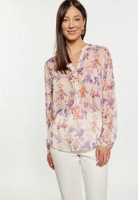 Blumenbluse aus leichtem Stoff, mit einem Mix aus rosa, lila und orange farbigen Mustern, Knöpfen an der Vorderseite und einem V-Ausschnitt. Kombiniert mit weißen Hosen.