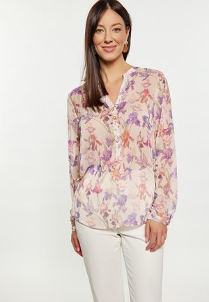 Blumenbluse aus leichtem Stoff, mit einem Mix aus rosa, lila und orange farbigen Mustern, Knöpfen an der Vorderseite und einem V-Ausschnitt. Kombiniert mit weißen Hosen.