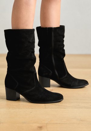 Jambes portant des bottes en daim noir mi-mollet avec des talons carrés et des fermetures éclair sur le côté, posées sur un sol en bois clair.