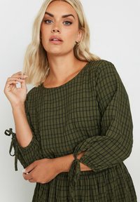 Vihreä ruudullinen mekko, jossa on teksturoitua kangasta, pitkät hihat, joissa on nauhat yksityiskohtina hihojen päissä, sekä pyöreä pääntie.