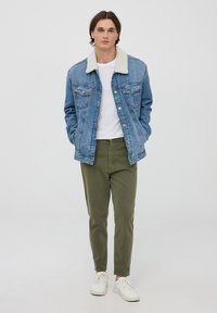 Giacca di jeans con colletto in pelliccia crema, di colore blu, con due tasche sul petto, abbinata a pantaloni verde oliva e scarpe da ginnastica bianche.