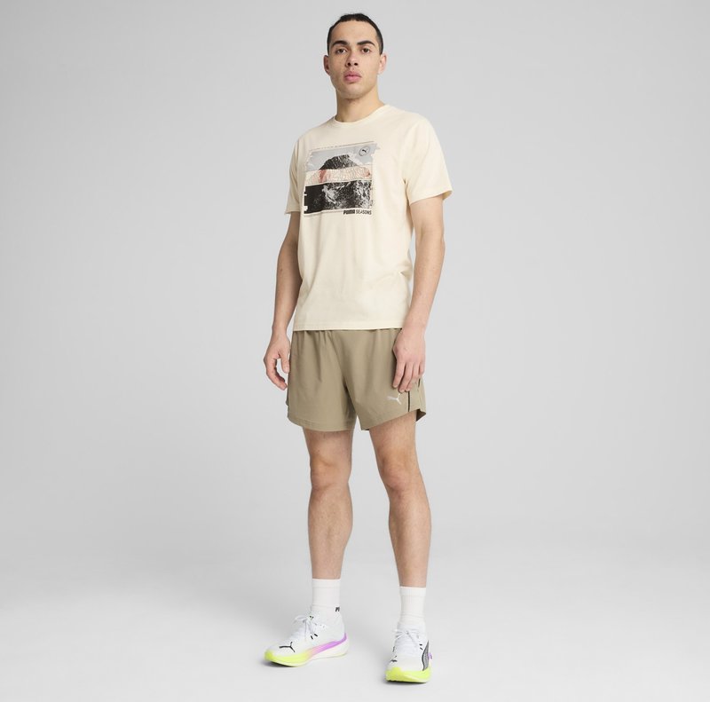 Lys beige t-shirt med grafisktryk, parret med khaki shorts. Modellen bærer hvide sneakers med gule detaljer og hvide ankelsokker.