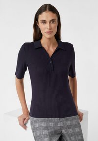 Granatowa koszulka polo w prążki z kołnierzykiem, krótkimi rękawami i zapięciem na guziki. Noszona z szarymi spodniami w kratę, o gładkiej fakturze.