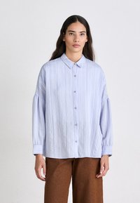 Lichtblauwe button-up blouse met verticale strepen, staande kraag en lange mouwen, gecombineerd met bruin wijde broekspijpen.