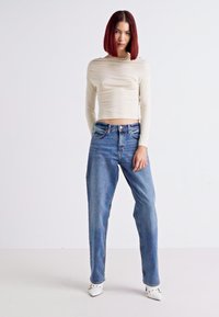 Crème, lang mouwtop met een aansluitend, gekreukt ontwerp; hooggetailleerde, lichtblauwe denim jeans; model draagt witte, puntige schoenen.