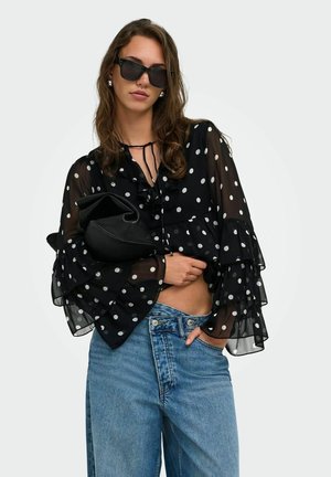Femme portant de grandes lunettes de soleil noires, un chemisier noir transparent à pois avec des manches à volants, un jean bleu clair, tenant une pochette noire.