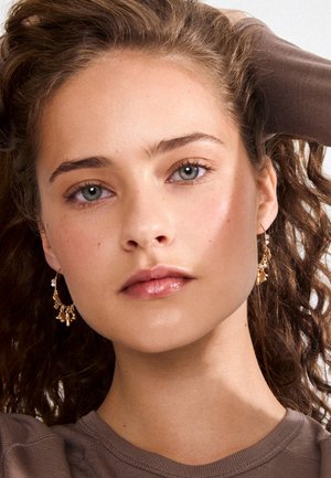 Mlada žena s kovrčavom smeđom kosom, nosi zlatne viseće naušnice i smeđu bluzu, gleda izravno s neutralnim izrazom.