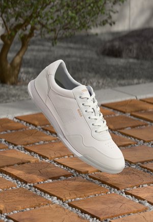 Baskets blanches en cuir avec lacets et logo BOSS flottant au-dessus d’un sol en carrelage en bois avec de petits cailloux blancs entre les carreaux.