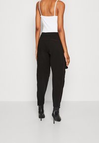 Černé cargo kalhoty s kopinatým střihem, elastickým pasem a detaily v červeném prošití. Kombinované s bílým tílkem a černými botami na podpatku.