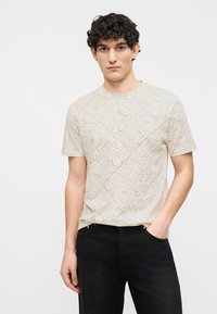 Νέος άνδρας με σγουρά μαλλιά που φοράει μπεζ T-shirt με σχέδια και μαύρο τζιν, στέκεται με το ένα χέρι στην τσέπη μπροστά σε απλό φόντο.