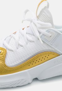 Baskets blanches avec maillage texturé et accents en cuir doré, comportant des lacets blancs, une semelle rembourrée et de petites perforations sur le côté.