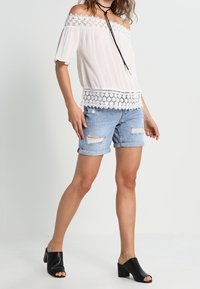 Vit blus med bar axel och spetskant, kombinerad med ljusblå slitna denimshorts. Svarta sandaletter med klack fulländar outfiten.