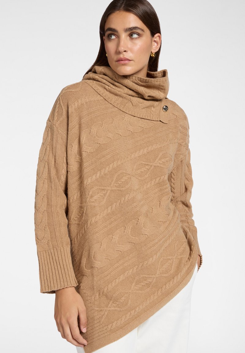 Kobieta ubrana w kamelowy asymetryczny sweter z przewlekanym splotem, o wysokim założonym kołnierzu i detalem w postaci guzika, w połączeniu z białymi spodniami.