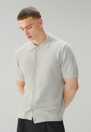 Noor mees lühikeste pruunkarvadega, seljas helehall lühikeste varrukatega nööbitav kootud särk ja mustad püksid, vaadates alla, käed selja taga.