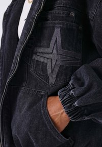 Zwarte denimjack met ritssluiting aan de voorkant, elastische manchetten en een zak met een grijs gestikt sterontwerp. Getextureerde stof met subtiele schaduwwerking.