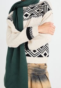 Personne portant un pull beige avec des motifs géométriques noirs, une écharpe en tricot verte et une jupe plissée beige délavée, les bras croisés sur le torse.