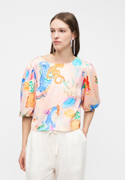 Femme portant un chemisier pastel à manches bouffantes avec un imprimé de créatures marines colorées, assorti à un pantalon blanc à cordon de serrage et des longues boucles d'oreilles en cristal.