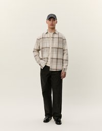Kariertes, beige Button-up-Flanellhemd mit braunem Karo, über einem hellbeigen Oberteil getragen; kombiniert mit dunklen, geraden Hosen und schwarzen Schuhen.