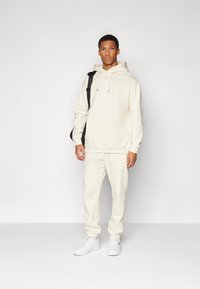 Kremfarget hettegenser og matchende joggebukser laget av mykt stoff, med en frontlomme og ribbestrikkede mansjetter. Brukt med hvite sneakers og en svart veske.