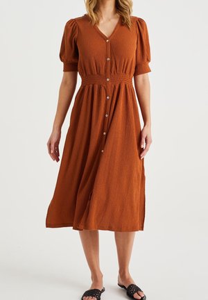 Vrouw draagt een roest-oranje midi-jurk met textuur, pofmouwen, knoopsluiting aan de voorkant, een elastische taille en zwarte geweven instapsandalen.