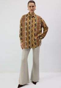 Chemise à manches longues avec un motif en zigzag vibrant jaune, marron et orange, associée à un pantalon évasé crème et des chaussures pointues foncées.