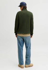 Pull en tricot vert avec un col montant, porté sur une chemise beige et un jean bleu, avec des poignets côtelés et un détail au dos.