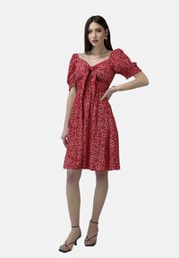 Rotes Blumenkleid mit Puffärmeln, einer Schnüroption vorne, knielangem Rock und leichter Struktur. Getragen mit schwarzen Sandalen mit Riemchen.