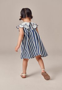 Blau-weiß gestreßtes Kleid mit gewelltem Kragen und Knopfdetail. Der Stoff scheint strukturiert zu sein. Kind trägt beigen Sandalen.