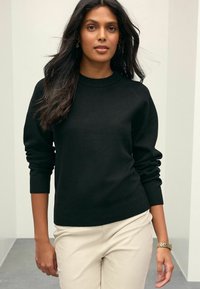 Femme aux longs cheveux noirs portant un pull noir, un pantalon beige, une montre en or et des boucles d'oreilles, se tenant à l'intérieur contre un mur à rayures claires.