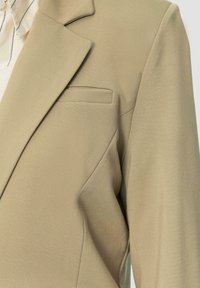 Beige op maat gemaakte blazer met geknipte revers, enkele borstzak en gestructureerde stof, met nadruk op strakke lijnen en een gestructureerd ontwerp.