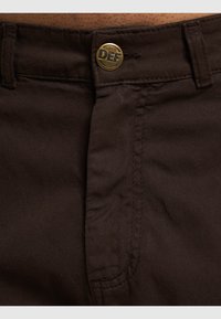 Pantalones de algodón marrón con una textura suave, que presentan un botón de metal grabado con "DEF" y costuras estándar a lo largo de las costuras.