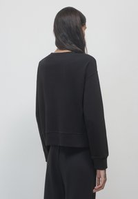 Felpa nera accorciata con maniche lunghe, caratterizzata da un collo arrotondato e polsini a coste, mostrata dal retro. Texture liscia.