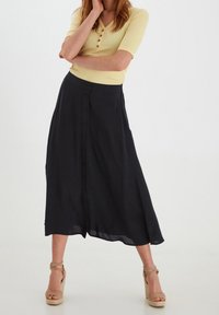 Černá midi sukně s plynulým designem, vybavená zapínáním na knoflíky vpředu a páskem v pase. Nošena s žlutým žebrovaným topem a béžovými lodičkami na platformě.