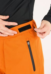 Oranger Outdoor-Hose aus strukturiertem Stoff. Verfügt über eine schwarze Reißverschlusstasche und einen Klettverschlussverstellriemen an der Taille.