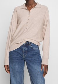 Maglione beige lavorato a maglia con colletto a polo, caratterizzato da una patta con bottoni e maniche lunghe. Abbinato a jeans di denim blu. Tessuto morbido.