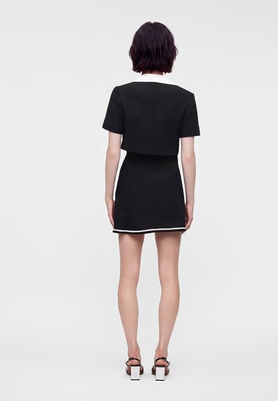 Femme aux cheveux courts et foncés portant une robe noire à manches courtes avec un col blanc et des bordures, ainsi que des sandales à talons blocs noires et blanches, debout de dos.