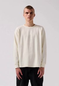 Giovane uomo con i capelli corti, che indossa una semplice camicia a maniche lunghe bianca e pantaloni neri, in piedi contro uno sfondo neutro.