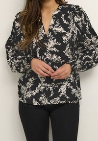 Blouse noire à motif floral blanc, encolure en V et manches longues bouffantes. Le tissu semble léger avec une texture lisse.